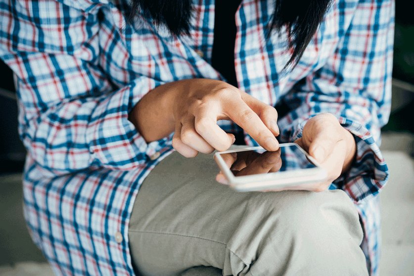A person sat down looking at their mobile phone wearing a checked shirt and brown trousers