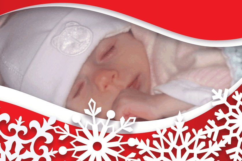 A baby in a white hat lies on their front with their thumb in their mouth the picture is in a red frame covered in white snowflakes