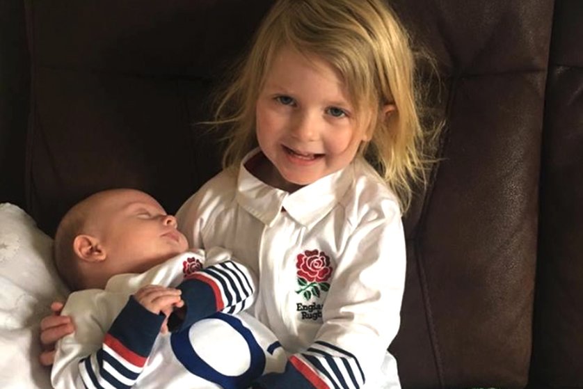 a small child with blonde hair in a white england rugby shirt smiles at the camera as a baby in an england rugby baby grow lies on her lap