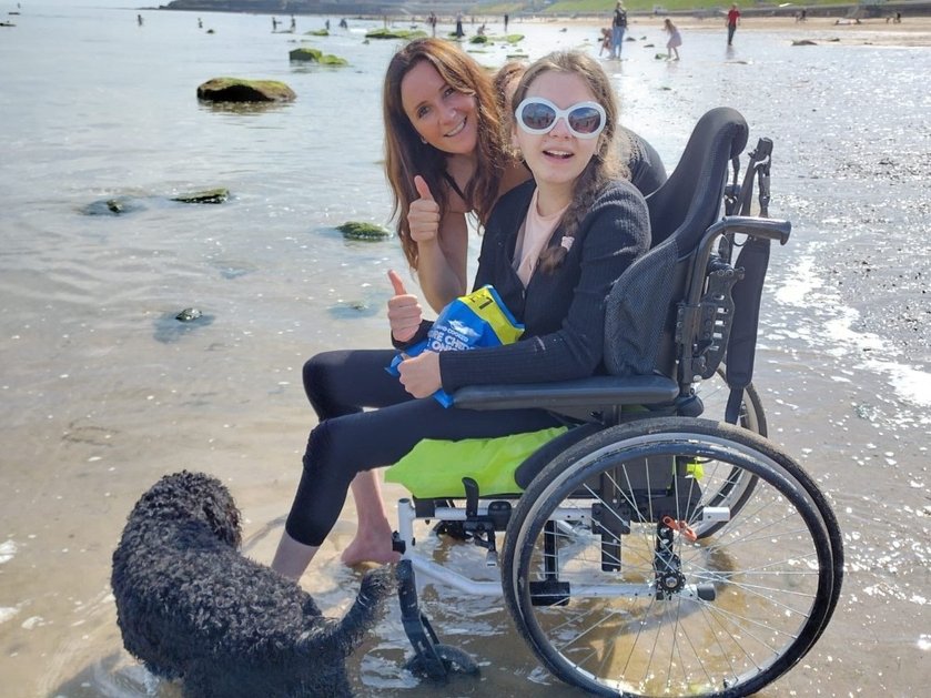 A young girl in a wheelchair at the edge of the sea with an older woman behind with her thumbs up