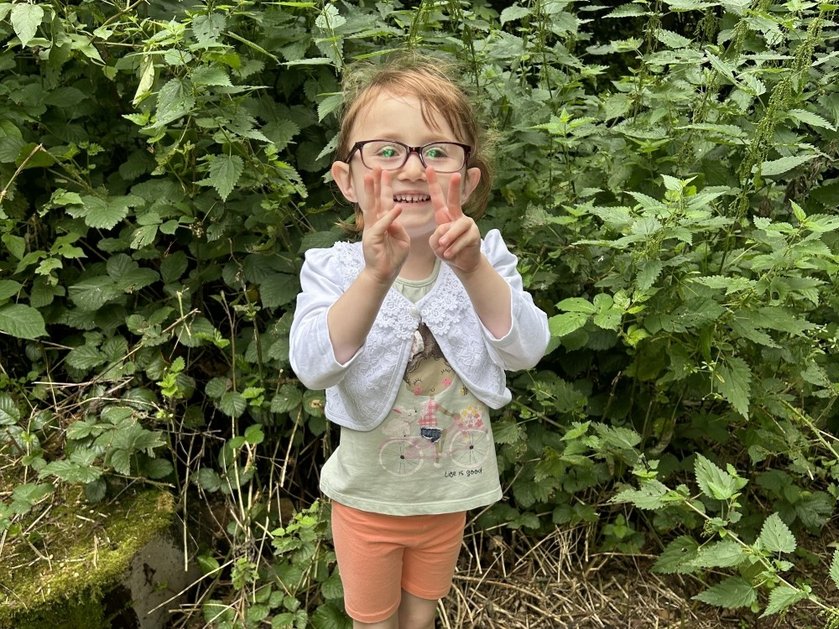 A young girl standing in front of a leafy hedge holding up her fingers and grinning