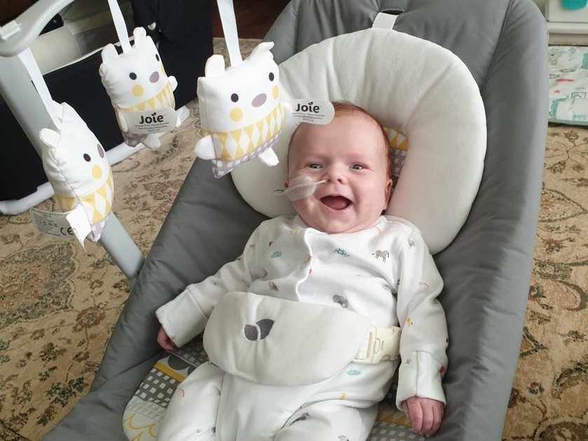 A baby in a seat laughing at the camera with a mobile above him