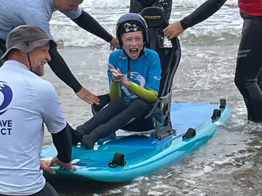 A young mito patient on a specially adapted surfboard laughing as he emerges from the sea