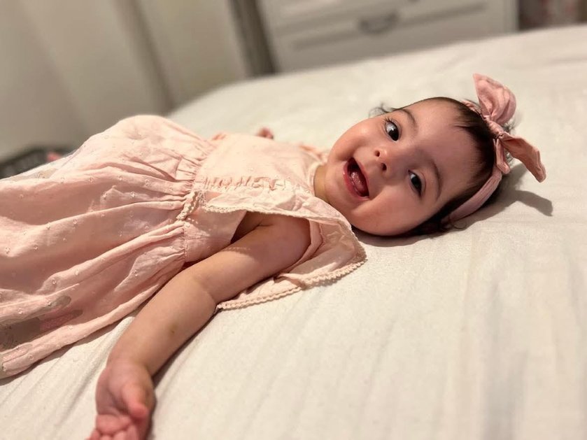 A baby in a pink dress lying down on a bed smiling