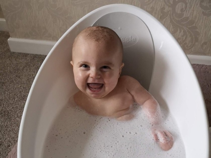 A baby in an oval bath looks up at the camera and laughs
