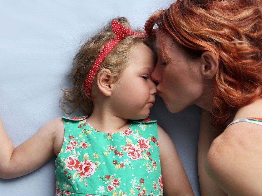 A woman with long red hair kisses a young girl who is lying down wearing a green floral dress