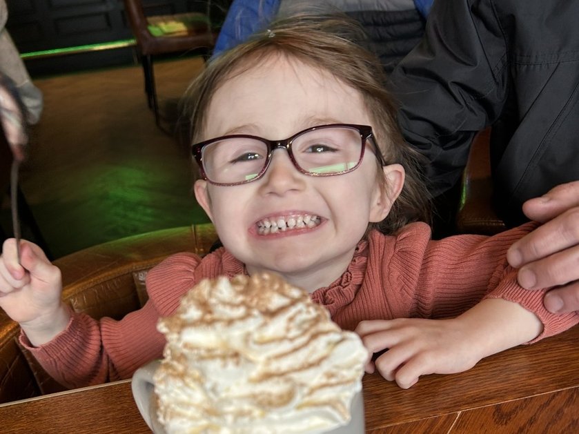 A young mito patient wearing glasses and grinning behind a large mug of hot chocolate topped with cream