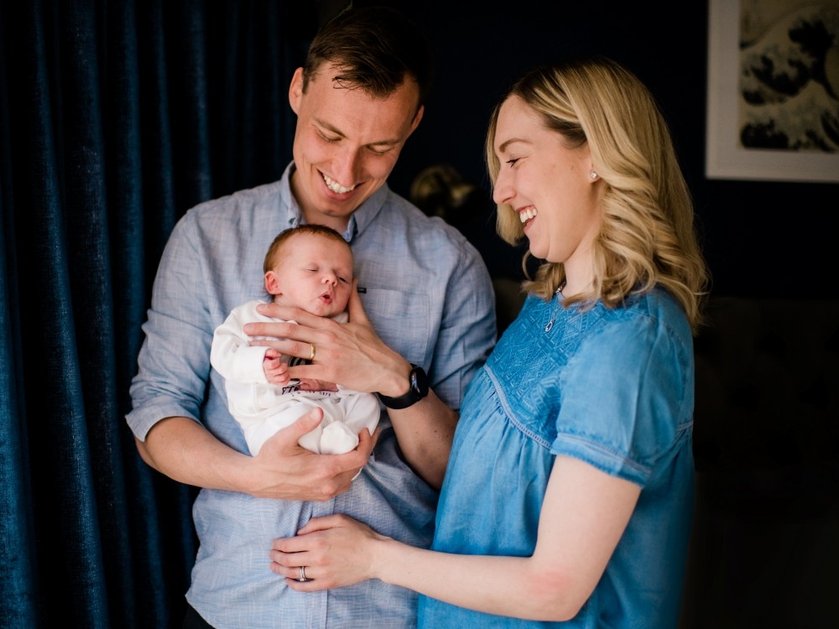A proud dad cradling a baby and looking down at him smiling with the mum standing next to them