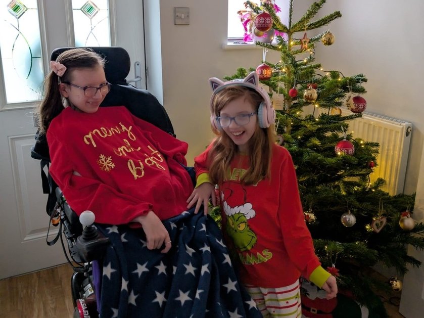 Two young siblings with mitochondrial disease together in a hall with a Christmas tree behind