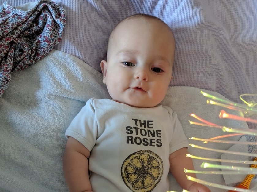 A baby lying on a bed wearing a Stone Roses babygro