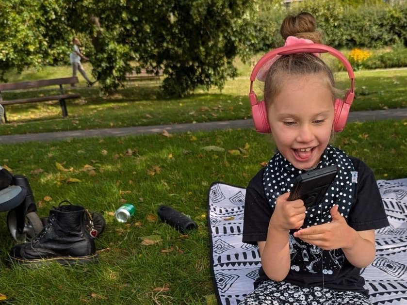 A young girl sitting in a park wearing pink headphones and smiling down at her phone