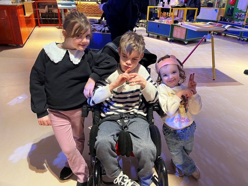 A young boy with mito sits in his wheelchair with his sisters standing either side smiling