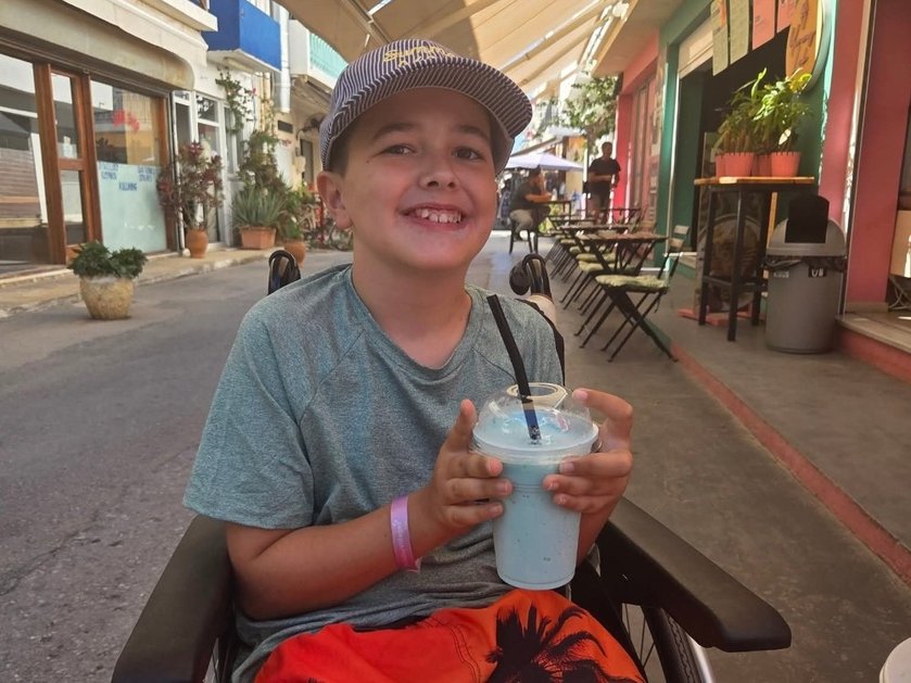 A young boy in a wheelchair wearing a baseball cap and holding a drink in a plastic cup with a straw