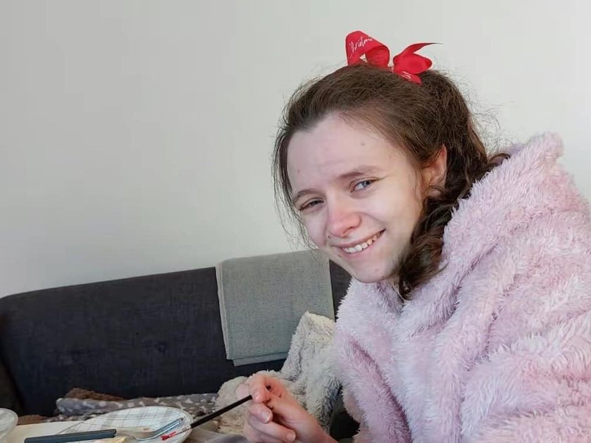 A young girl in a pink fluffy top and red hairbow smiling at the camera with a paintbrush in her hand