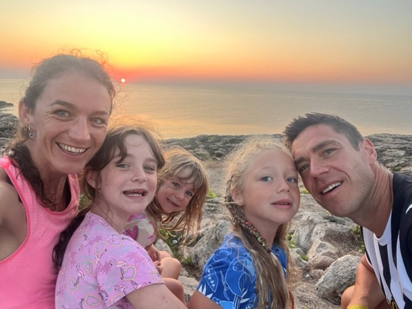 A mum, dad and three children smile at the camera with a coastal sunset behind