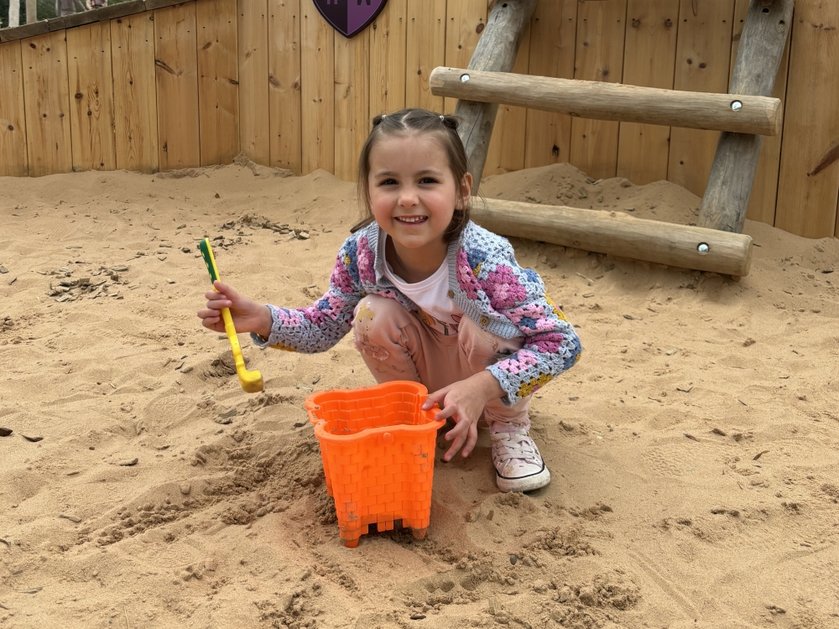 A young girl with her hair tied back playing in a sandpit with a bucket and spade