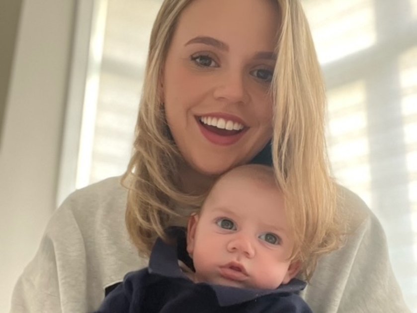 A lady with long blonde hair laughing and holding a young baby in front of her