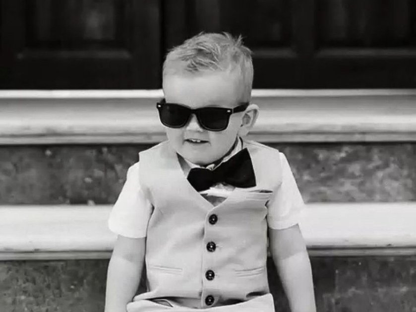Black and white image of a toddler wearing shades, a bow tie and waistcoat