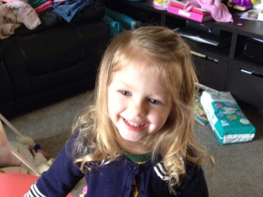 A young girl with long blonde hair smiling and standing with toys behind her