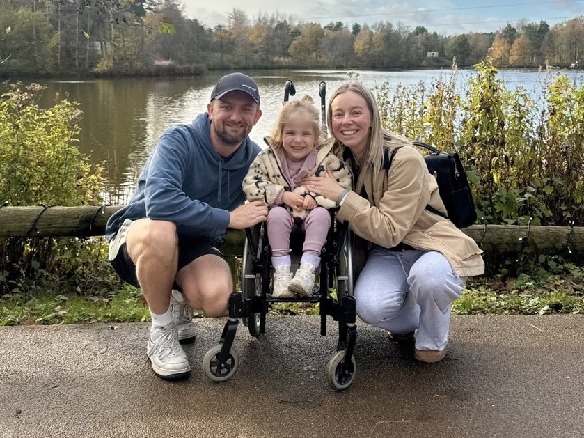 A smiling mum and dad crouch next to their daughter in a buggy in front of a lake