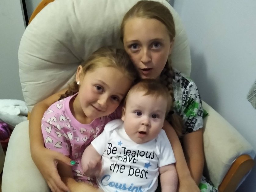 A baby mito patient sitting on a chair being cuddled by two older girls