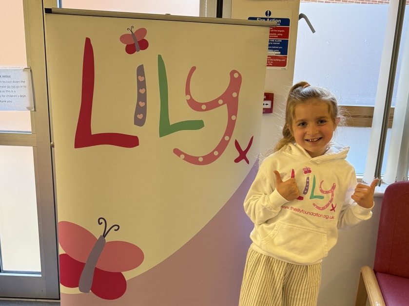 A young girl wearing a Lily hoodie standing in front of a large Lily banner with her thumbs up