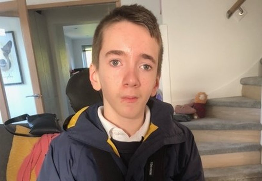 A boy with mitochondrail disease looking into the camera taken in a home with stairs in the background