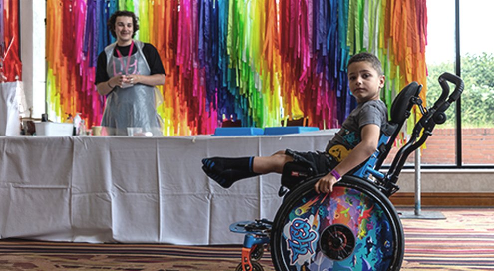 Young boy with mito, doing a wheelie in his wheelchair past a slime making station at family weekend.