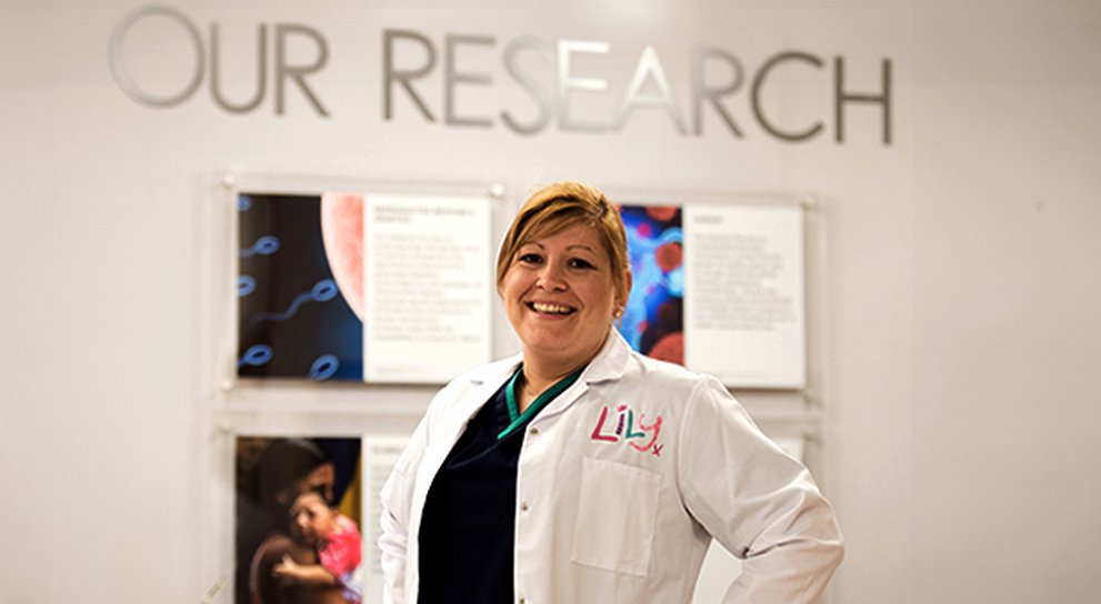Lady in a white lab coat with the Lily logo on top pocket, standing in front of a sign on the wall that says " Our Research"