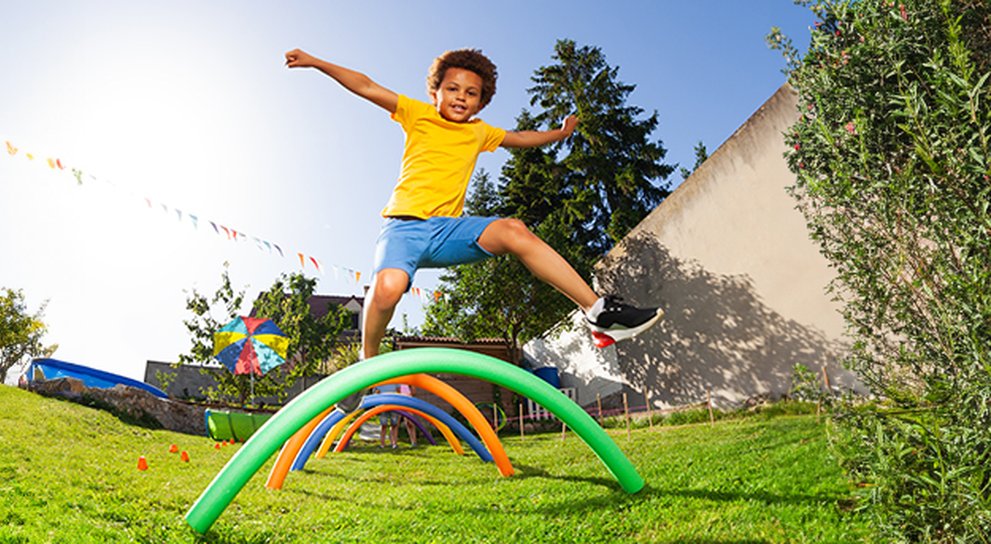 A sunny garden with colourful pool noodles stuck into the ground to make arches. A young boy is leaping over the hurdles