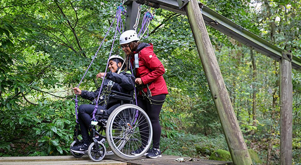 Young adult in her wheelchair being pushed by her mum over a forest assault course