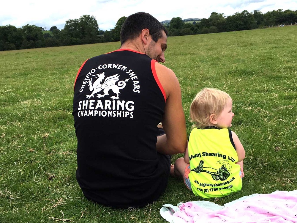 A man with his back to the camera in a black vests sits next to a small child in a yellow vest in a field