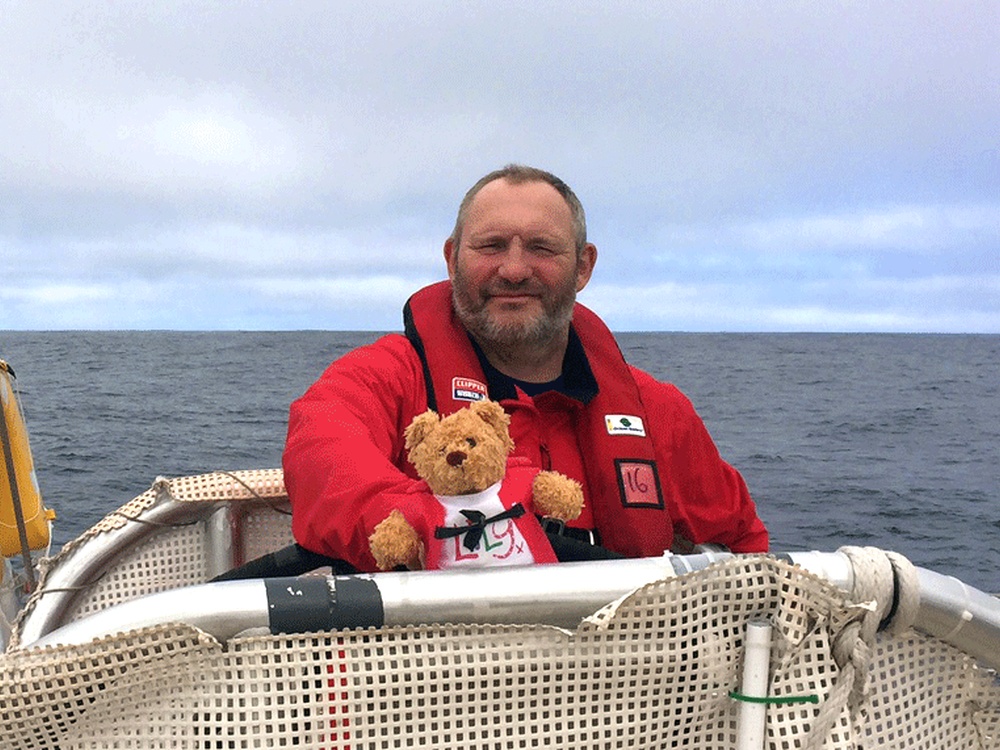 A man in a red waterproof sits holding a teddy bear with the ocean behind him