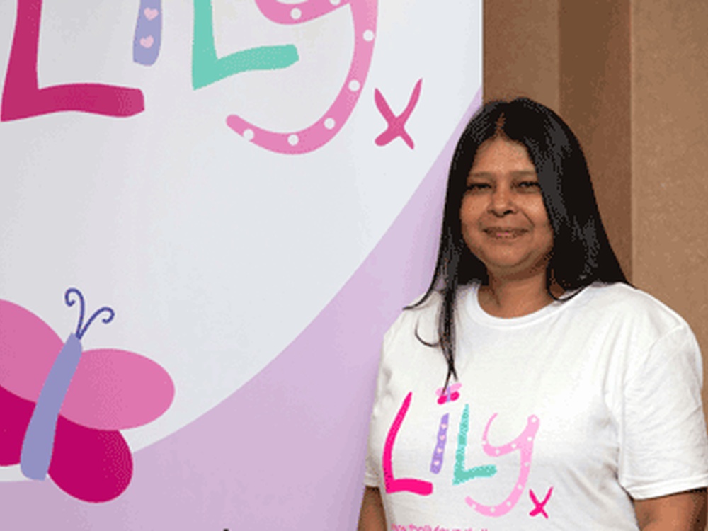 A women with long dark hair stands in front of a Lily Foundation bannor wearing a white lily foundation tshirt