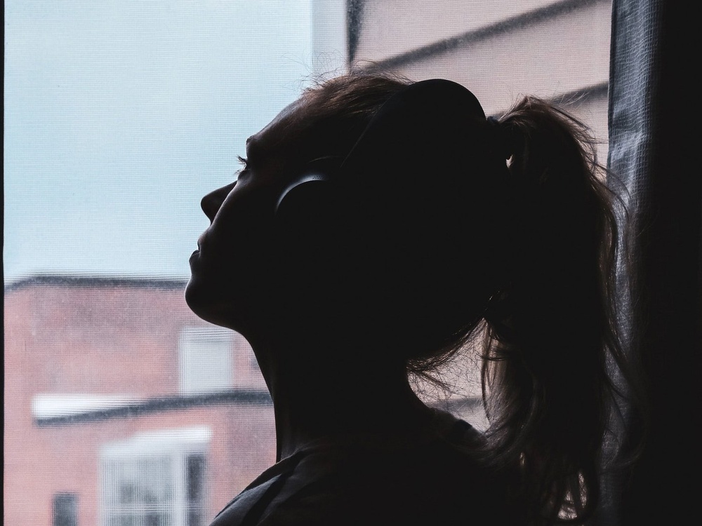 silhouette of a girl with her hair tied in a pony tail with head phones on, gazing up to the sky out of a window