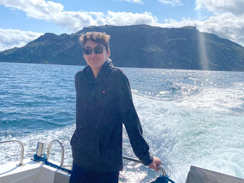 JoJo standing on the back of a boat, wearing sunglasses, with the waves of the sea and blue sky and hills in the background