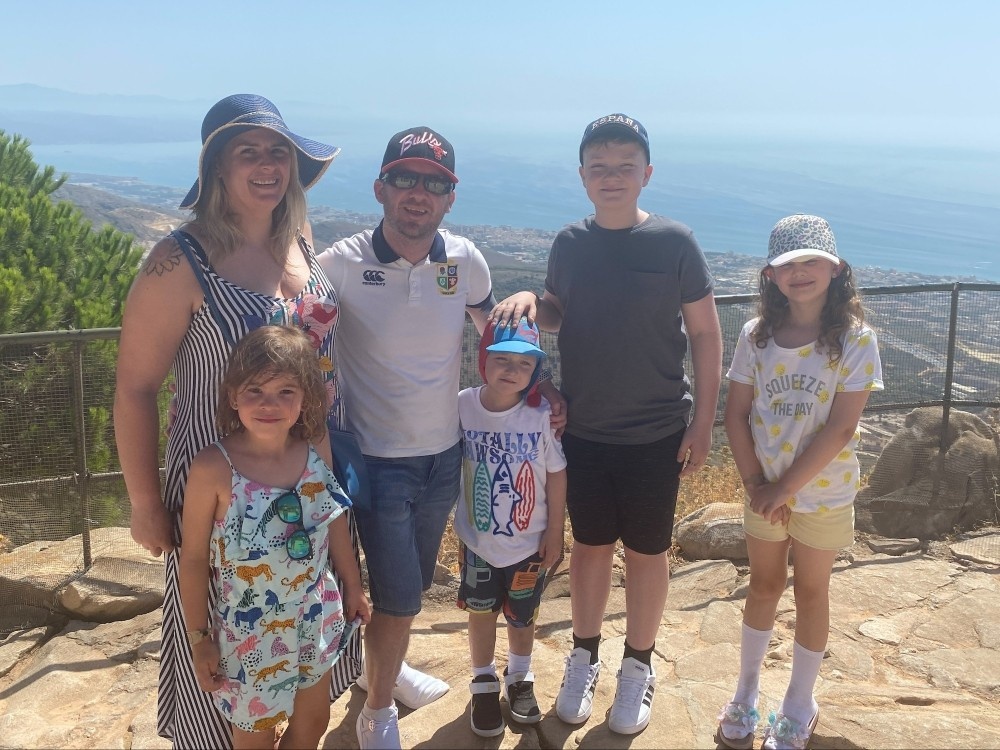 A family group stand together up on a cliff with land, sea and blue sky behind them