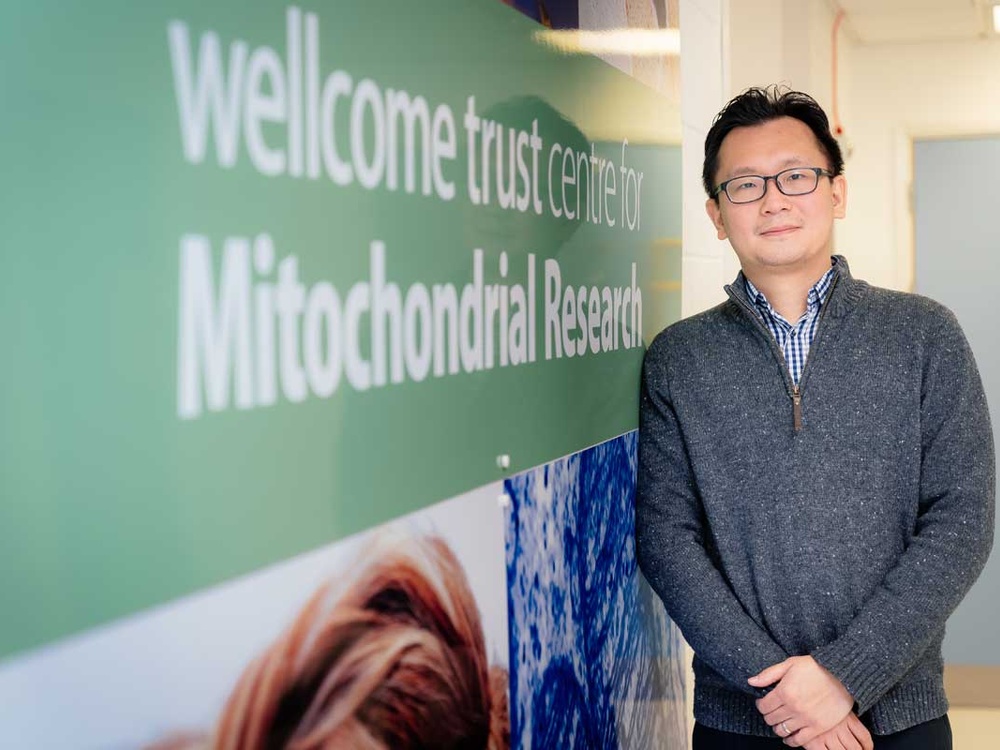 A man in glasses stands infront of a sign that says Welllcome Trust and Mitochondrial Disease