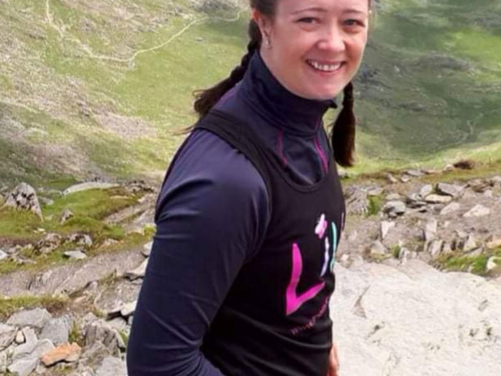 A women with dark hair in plaits in a lily jumper stand on the edge if a mountain