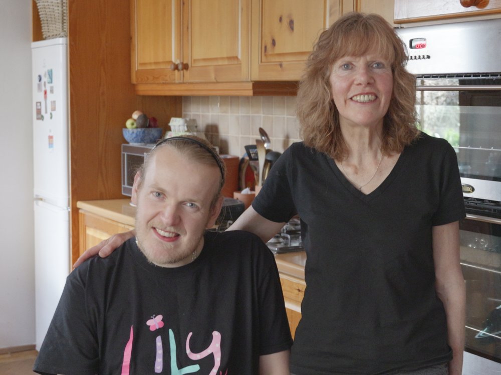 A woman standing with her arm around a young man in a Lily top in a wheelchair in their kitchen