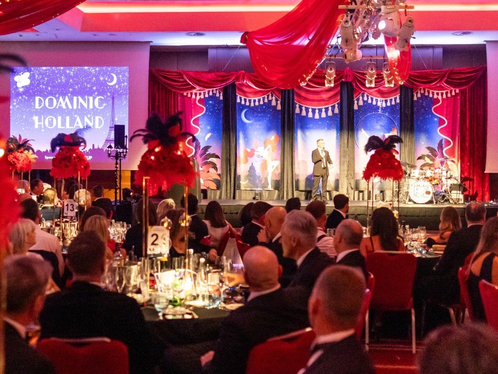 Lily ball host Dominic Holland on stage in front of several tables of people in black tie outfits