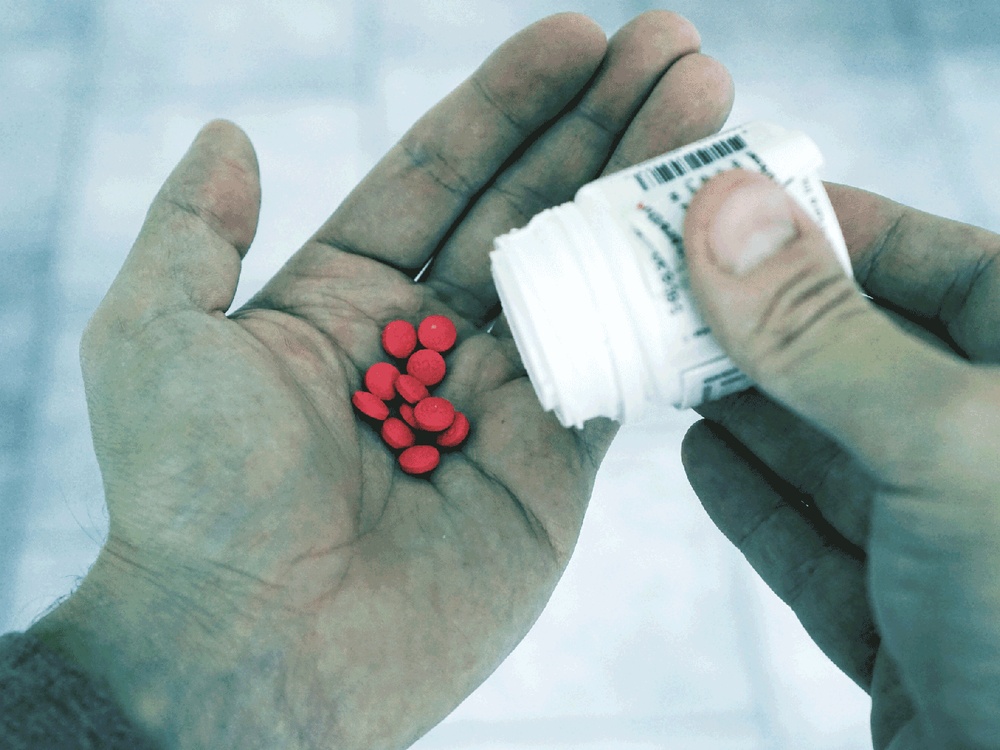 An open hand with red coloured tablets in the palm being poored out of a white pill bottle by the other hand