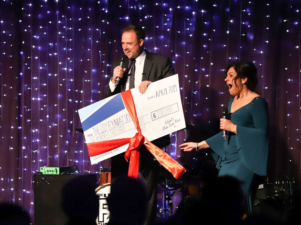 A man on a stage holding a microphone and a large cheque next to him is woman with dark hair and a green dress