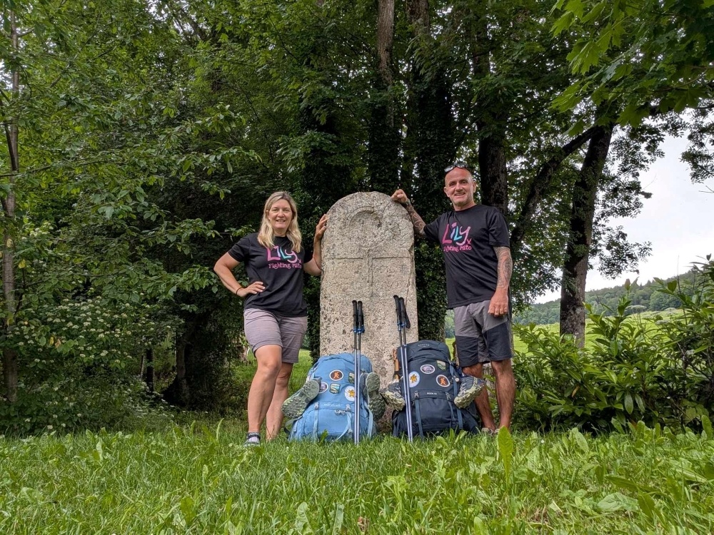 A couple wearing Lily Foundation tops and standing either side of a stone monument with their hiking rucksacks