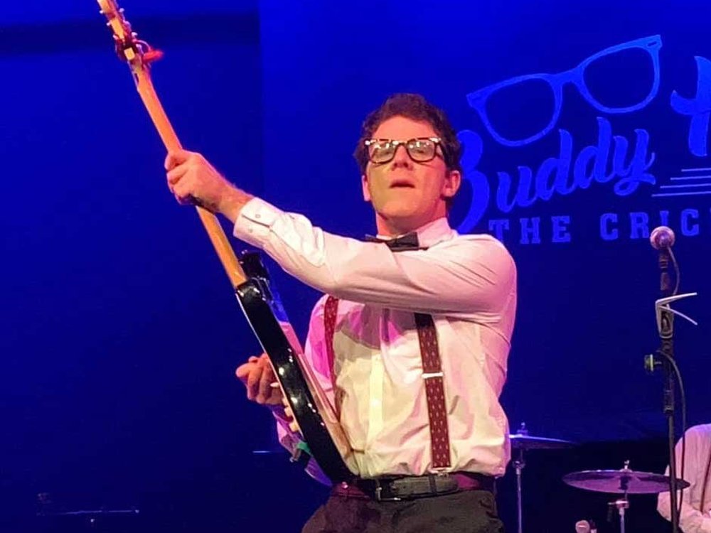 A man wearing glasses wearing a white shirt and braces holding a guitar