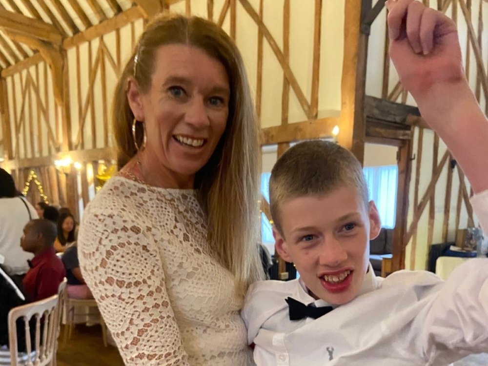A lady in a cream dress laughing and holding a young man with mitochondrial disease