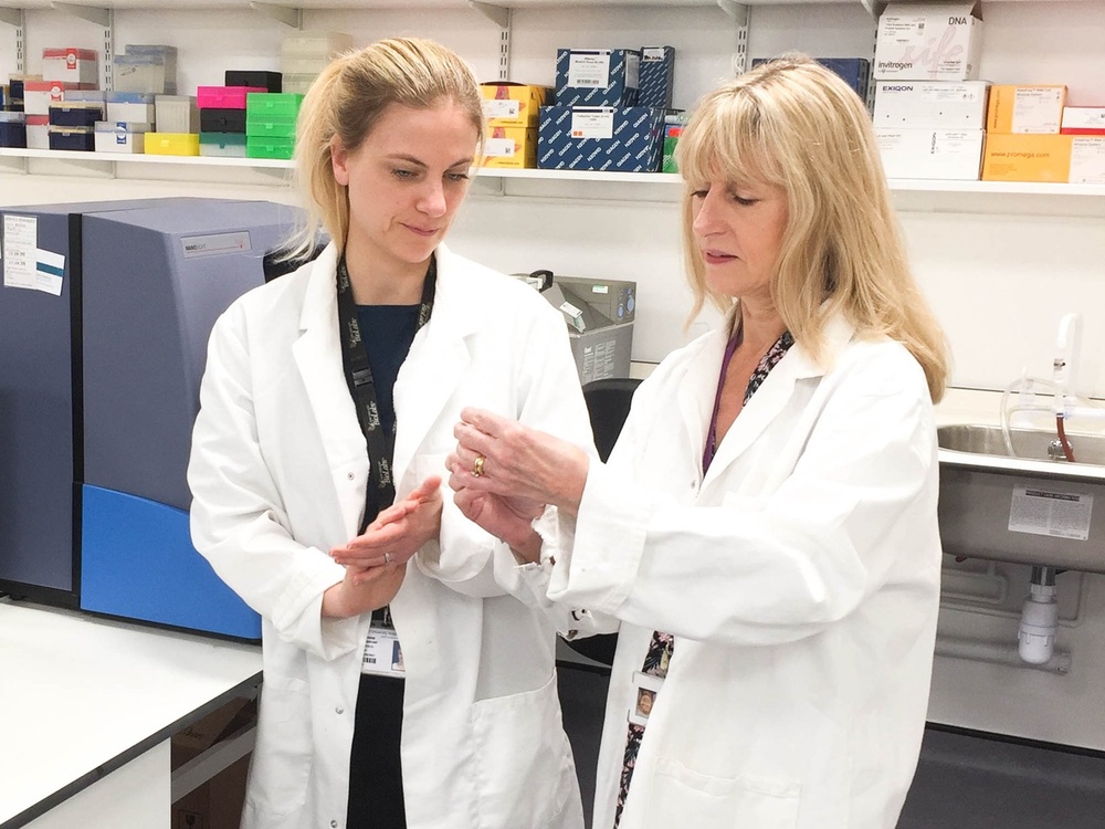 Two women in white lab coats stand in a laboratory. One is holding something up in her hands and the other is looking at it