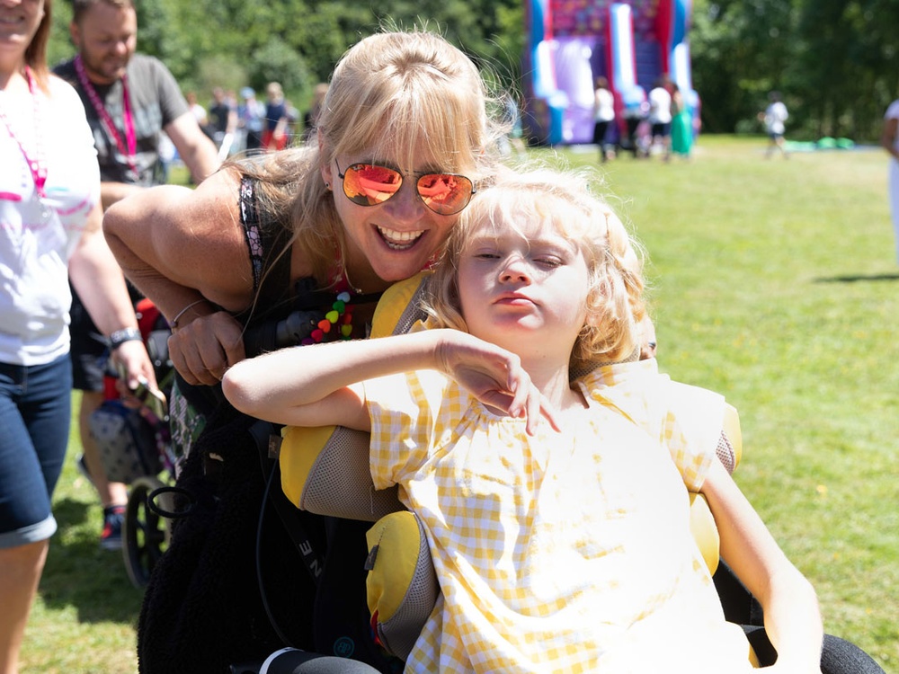 A women in sunglasses smiles at the camera pushing a girl in a wheelchair