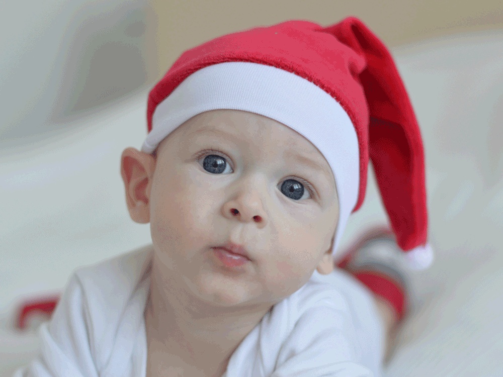 A head shot of a baby with blue eyes in a red Santa hat