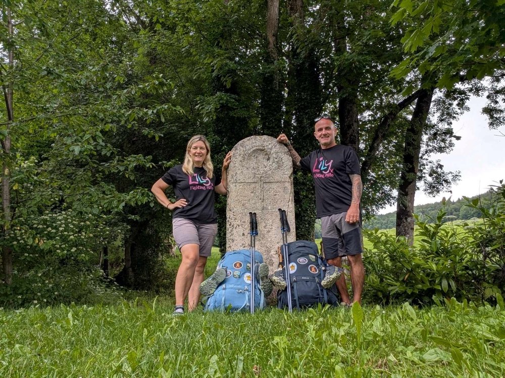 A couple wearing Lily Foundation tops and standing either side of a stone monument with their hiking rucksacks